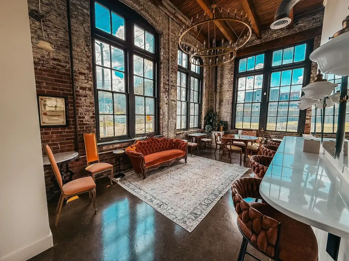 Wide shot of the Rosewood Room bridal suite at 24 Shelby with exposed brick wall, huge arched windows flooding daylight, a leather chesterfield sofa, vintage rug, and copper chandelier. The natural-light getting-ready space the venue includes with every booking
