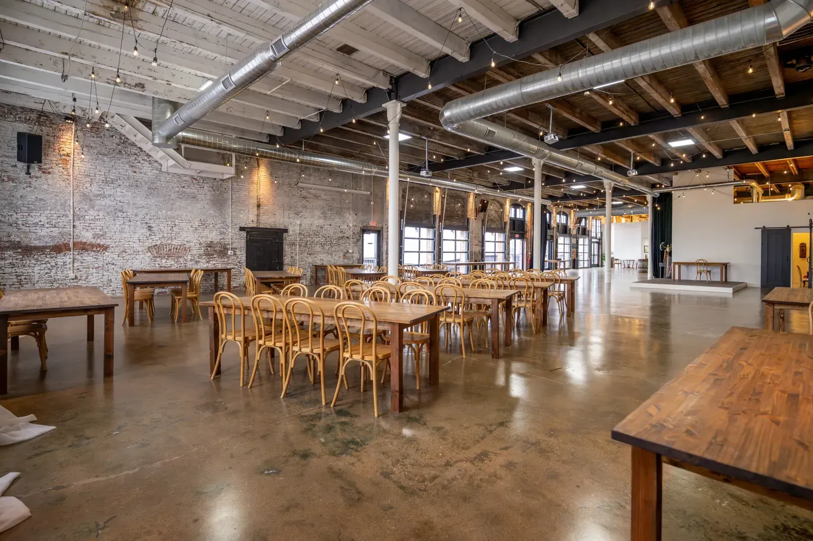 The Edison Room at 24 Shelby set with long wooden tables and bentwood chairs under Edison string lights and original brick, the blank canvas for a black and gold year-end gala