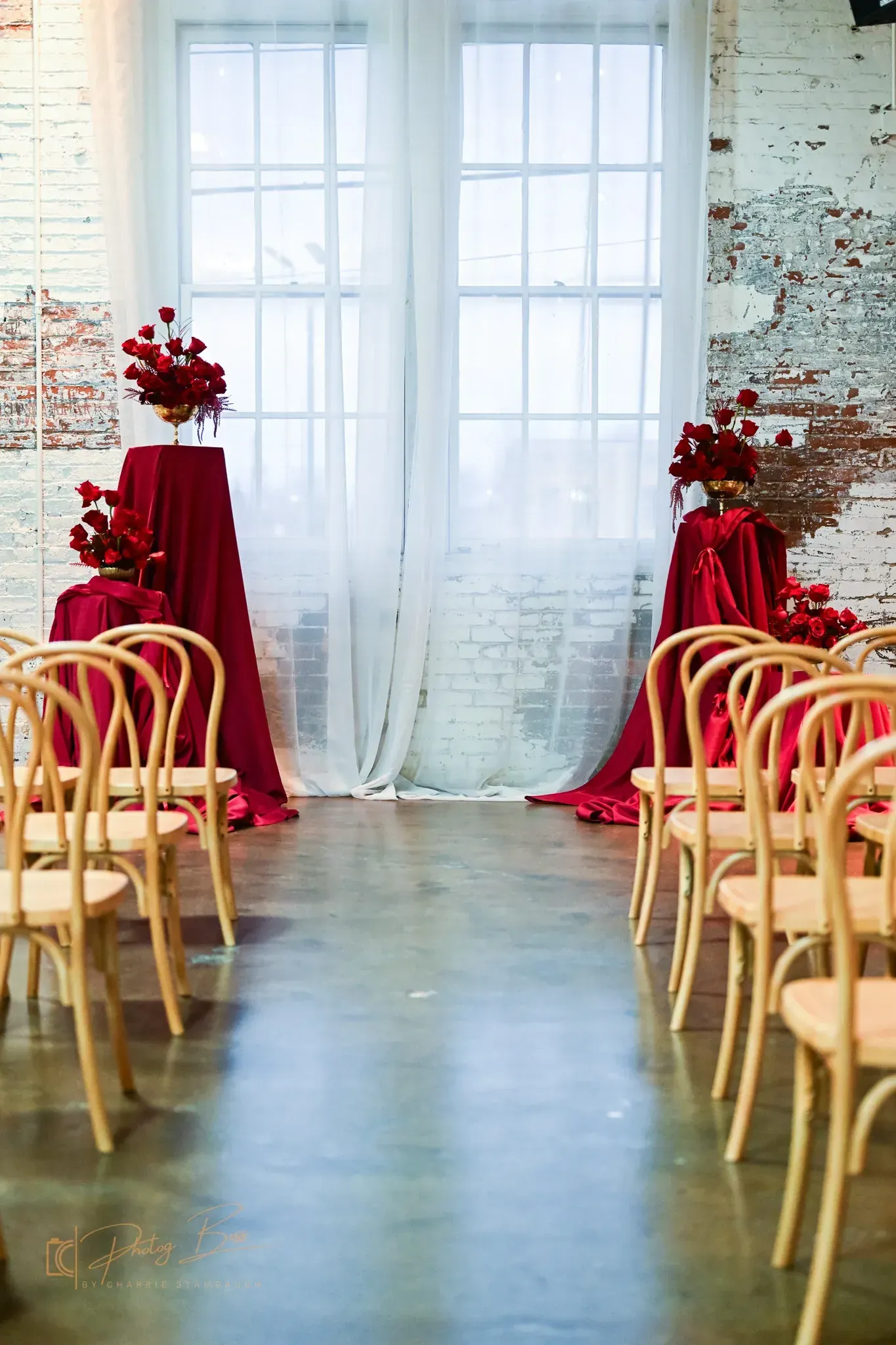 Indoor ceremony at 24 Shelby for an Indianapolis wedding in 2026, with rows of light wood chairs facing an altar of bold red drapery and oversized red and gold floral arrangements against original 1898 brick walls.
