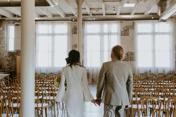 Couple walking hand in hand down the ceremony aisle at 24 Shelby, flanked by bentwood cross-back chairs and a 'Choose a Seat, Not a Side' welcome sign, inside the historic 1898 venue with original industrial trusses and oversized windows. One of the most unique wedding venues in Indianapolis.
