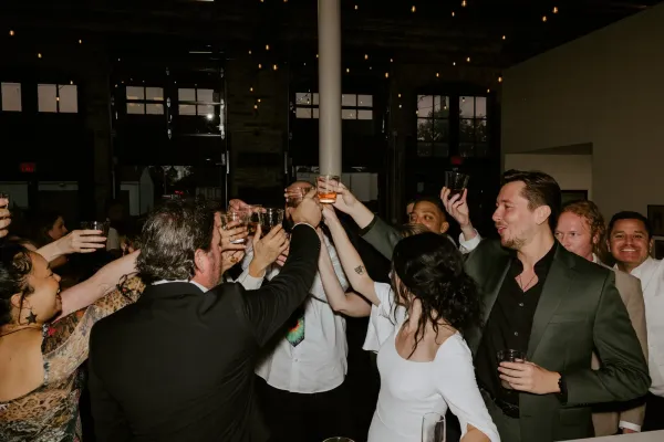 Guests raising a toast under Edison string lights at a holiday party at 24 Shelby, a historic downtown Indianapolis venue, with original 1898 brick and oversized windows behind them.