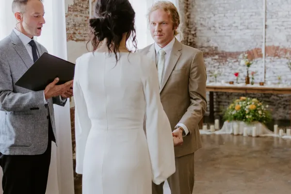 Intimate wedding ceremony at 24 Shelby in downtown Indianapolis with the bride and officiant standing in a softly lit historic space, exposed brick walls and Edison string lights overhead, set up for a small celebration of fewer than 75 guests.