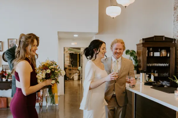 Three bridal shower guests toasting cocktails at the Libations Lounge bar inside 24 Shelby in downtown Indianapolis. Bride in white, bridesmaid with a bouquet, and a guest raise glasses against the bar back, exposed brick, and warm natural light.