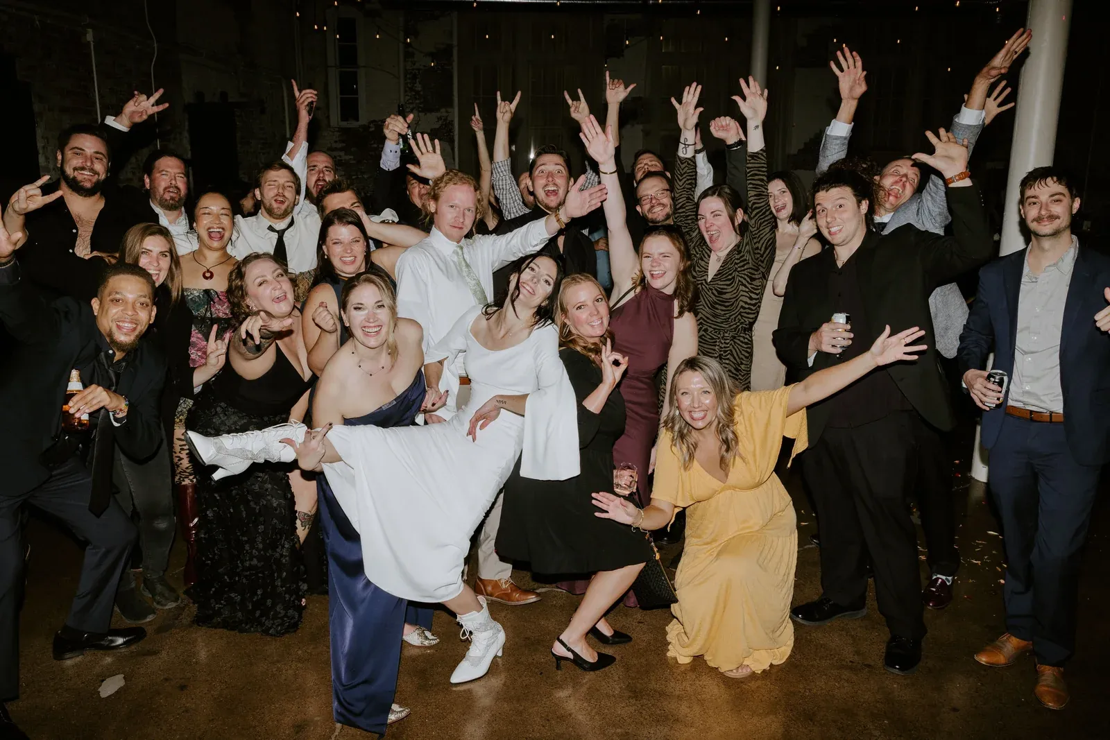 A large group of guests cheering with arms raised at a party inside 24 Shelby, a downtown Indianapolis birthday and graduation party venue, under Edison string lights and exposed brick.