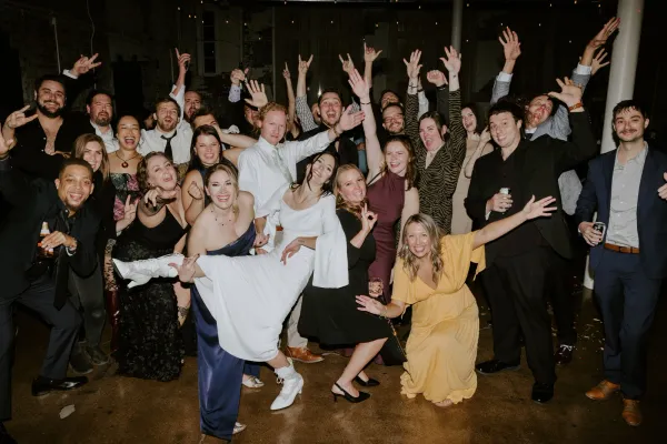 A large group of guests cheering with arms raised at a party inside 24 Shelby, a downtown Indianapolis birthday and graduation party venue, under Edison string lights and exposed brick.